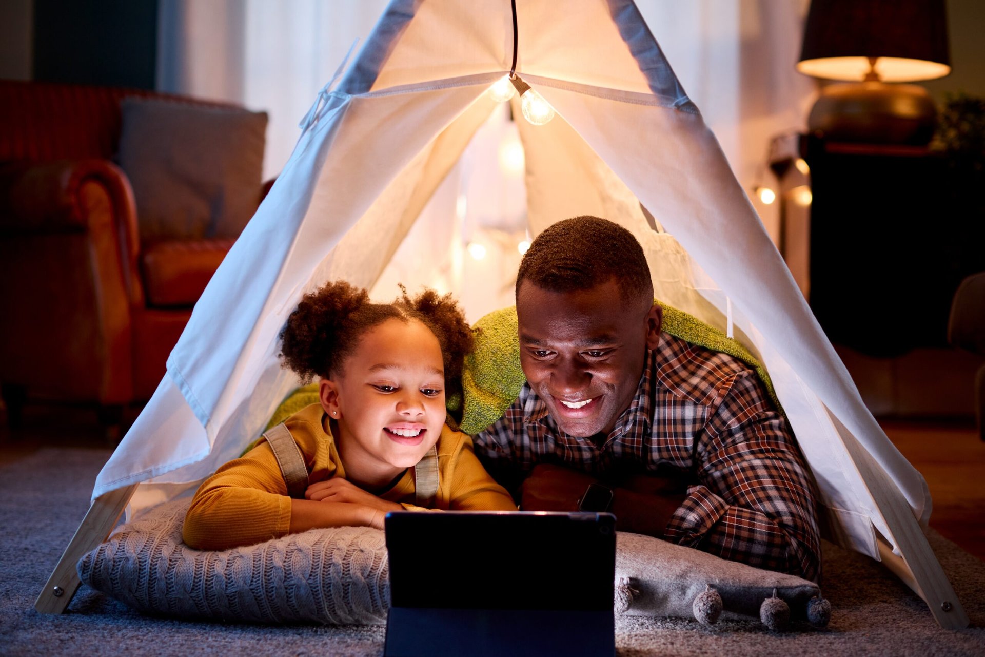 Father and daughter at home lying in an indoor tent streaming content on a tablet